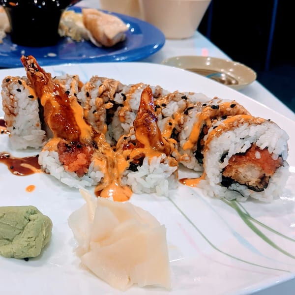 Sushi rolls with shrimp tempura, spicy sauce, sesame seeds, wasabi, and ginger on a white plate.