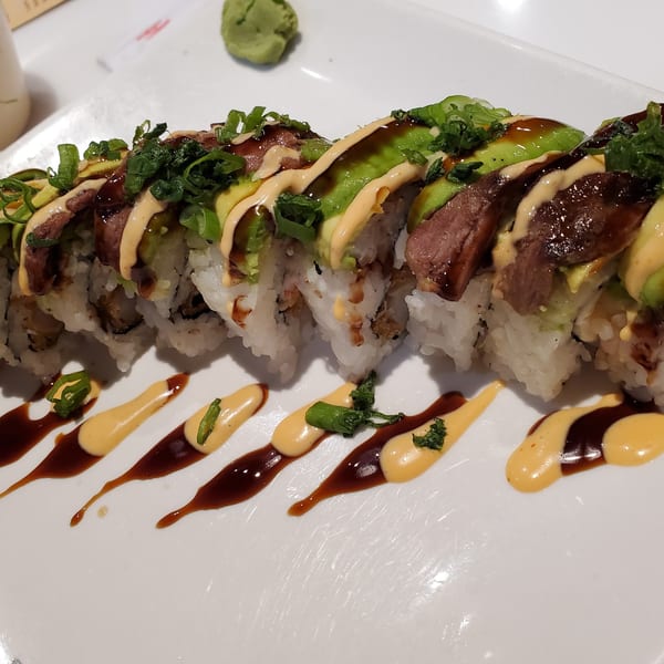 Asian-fusion sushi roll with beef, avocado, green onions, and two sauces on a white plate.
