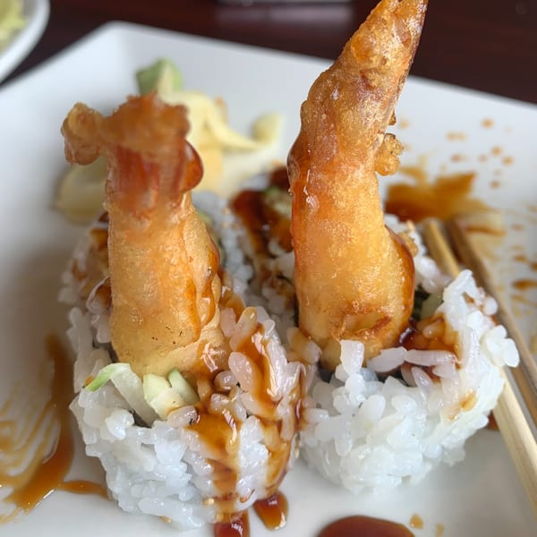 Two pieces of Lim Dynasty Asia Cafe & Xpress tempura shrimp sushi rolls with sauce and chopsticks.